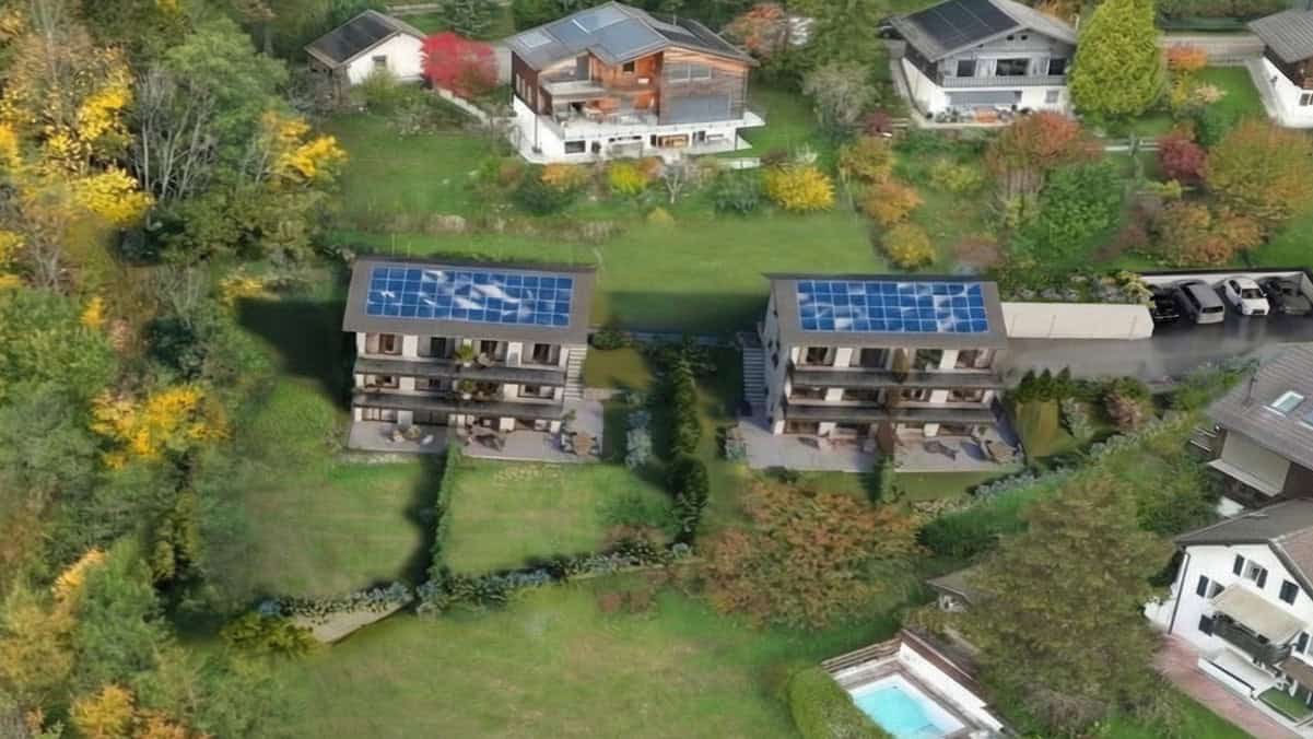Vue aérienne d’un petit ensemble résidentiel de deux immeubles neufs à toits équipés de panneaux solaires, entourés de jardins, d’arbres et de villas voisines.